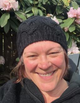 Close up of Ellen smiling at the camera. She has pale skin, chin-length hair and a bright smile. She is wearing a black beanie and black sweater.